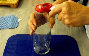 A tall glass gar jar sits on top of a blue placemat. One hand holds the neck of the jar while another hand holds a red jar opener at the edge of the lid. 