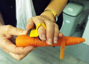 A person holds a carrot horizontally at one end. A yellow ring of a palm peeler is attached to the index finger of the other hand on top of the carrot.