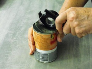 A can of tomatoes sits on a bench. One hand grips the can while another holds a ring pull opener with plastic hook. The ring is lifting and pulled to peel back lid.