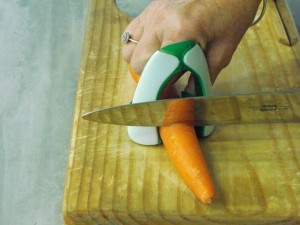 A carrot sits on a wooden chopping board. A hand holds the end of the carrot in place, with a safe slice in between the hand and a knife chopping the carrot. 