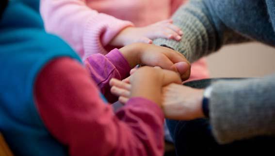 One child lays their hands over and grasps the hands of an adult. Another child rests their right hand on the adult's forearm. Only the arms and hands are visible in the photo