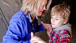 A woman in a purple jacket is face to face with a child wearing a checked shirt. The woman has her hands under the outspread hands of the child. She is using tactile signing