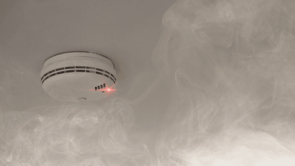 a white round smoke alarm is attached to a ceiling. It has a flashing red light and is surrounded by smoke