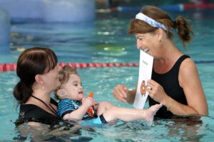A woman standing in a swimming pool,  holds a young boy with his back into her chest. He is holding an orange toy fish. They are looking at a woman who is gesturing with her right hand, imitating the movement of a fish and holding a picture of an orange fish in her left hand. They are smiling at each other.