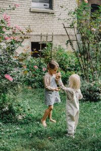 Two children are outside among rose bushes. One child has her thumb in her mouth, while the other child holds onto this hand.