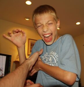 A young boy is laughing. His right hand is raised to his shoulder. His left hand is under his armpit and an adult's hand is is also in contact with this hand.