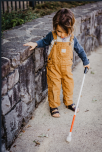 A young boy with brown overalls is holding a white cane in his left hand. With his right hand, he is holding onto a stone wall. The wall's height goes up to just below his shoulders. He is walking and looking towards the ground.