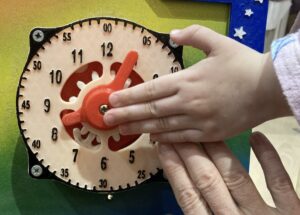 An adult's hand guides a child's hand towards a plastic clock. The child's hand is resting on top of the adult's fingertips.
