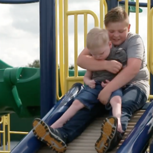 Two boys on a playground, going down a slide. The smaller boy is sitting on the other boy's lap. The other boy is supporting him to go down the slide.